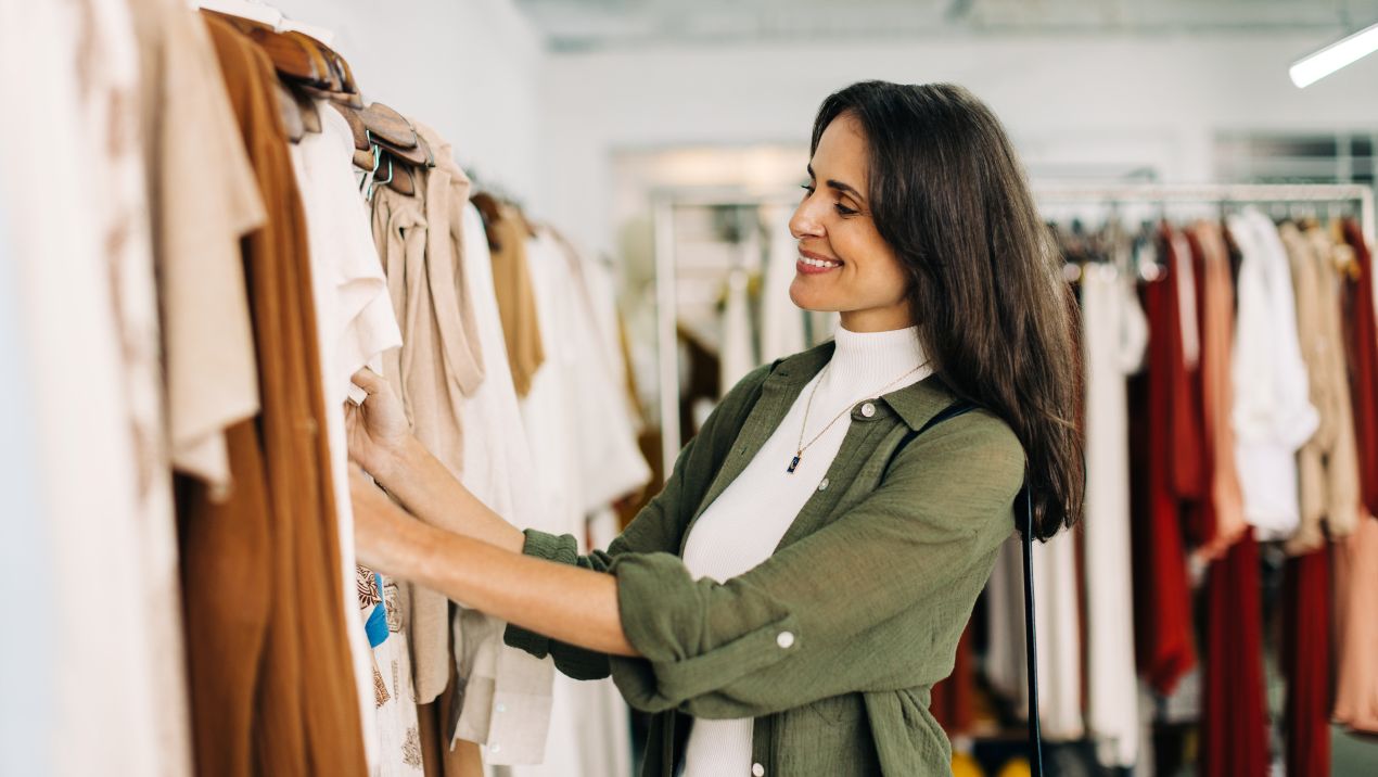 Frau beim Kleider kaufen, sieht sich beige Kleidungsstuecke in einer Boutique an